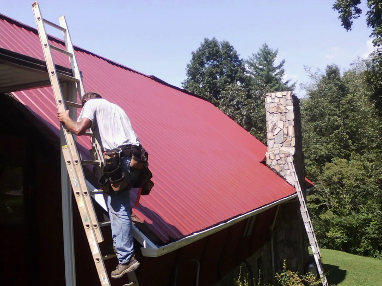 Roofer on a ladder beside a red metal roof and stone chimney — Soffit & Fascia Repair by Roofing Brothers in Walsh